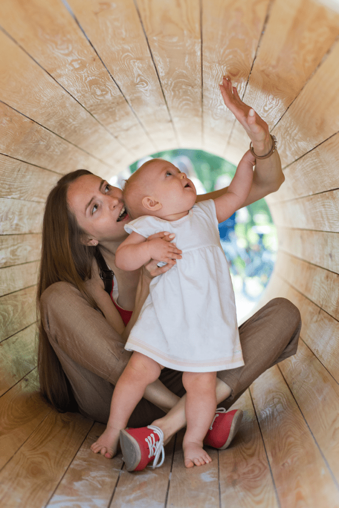A parent holds their baby while they explore the inside of a wooden tunnel at the playground.