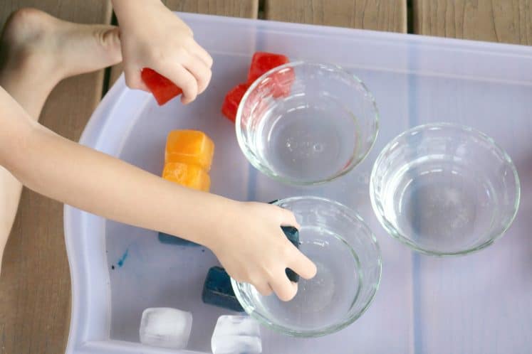 preschooler dropping colored ice cubes into clear dish filled with water