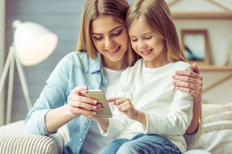  young mom and her little daughter are using a smartphone and smiling while sitting on sofa at home
