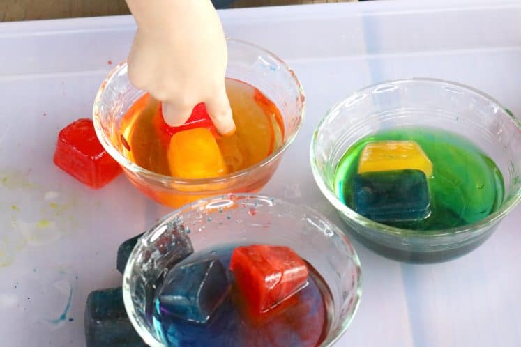 preschooler adding red ice cube to dish containing yellow ice cube to make orange colored water