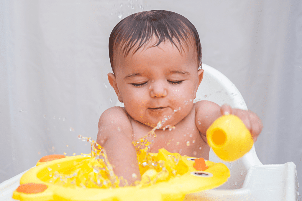 A baby sits in a highchair with their eyes closed. Their arms are outstretched, splashing water from a yellow tray in front of them. The baby is learning about sensory experience of water and listening to the splashes.