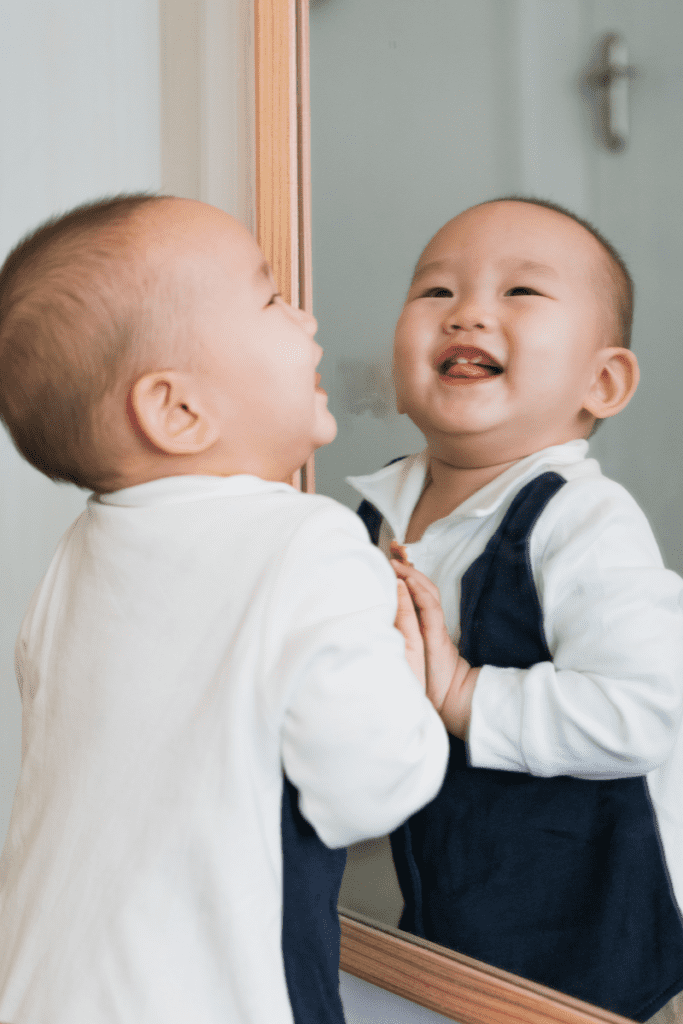 A baby who pulled themselves to stand is smiling into a mirror and exploring their reflection during a sensory experience.